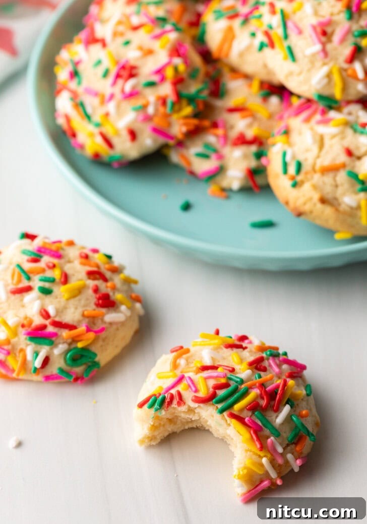 Cookie with sprinkles and a bit taken out is close to camera. Another cookie with no bite is off to the side. In the background is a plate piled up with cream cheese cookies.
