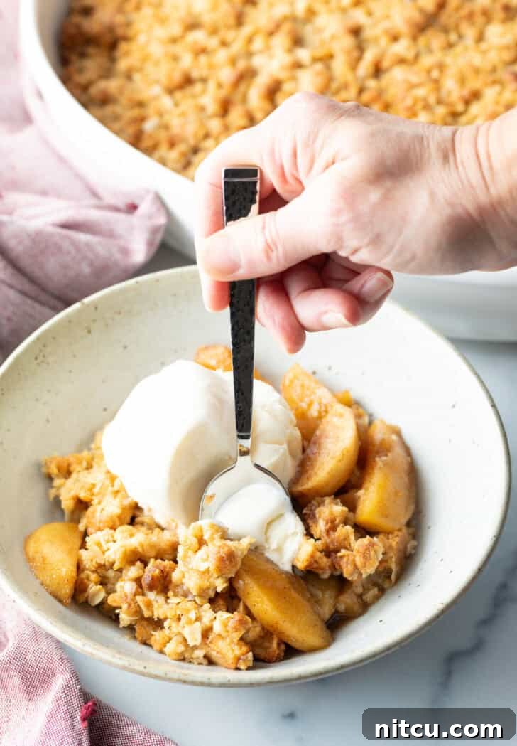 White bowl with golden apple crisp topped with a big scoop of ice cream. A hand with a metal spoon taking a bite of ice cream, showing pure enjoyment.