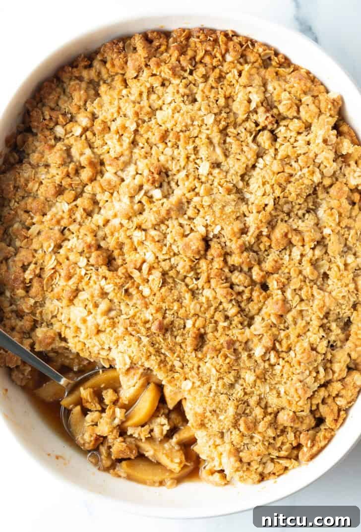 Top down view of baked apple crisp with oats. A spoon is scooping away a serving, showing the gooey apples and crunchy topping.