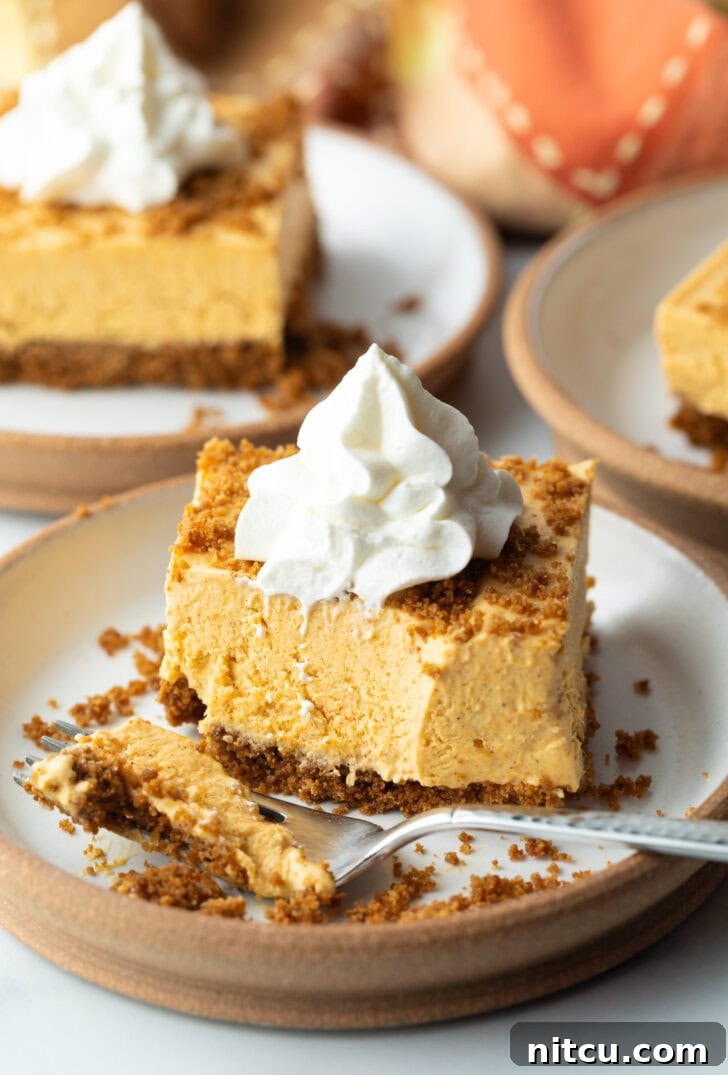 Square of pumpkin pie cheesecake on a white plate, topped with whipped cream. A fork with a scoop of the dessert is on the plate as well.