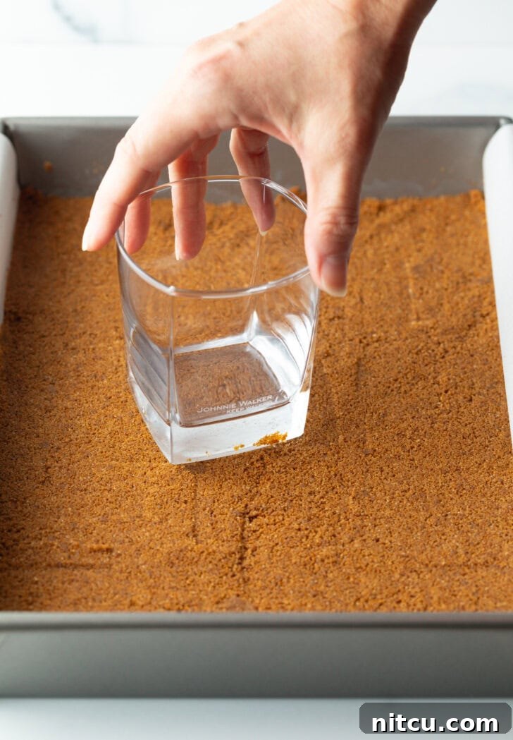 Hand with glass pressing the cookie base into the bottom of a baking dish.