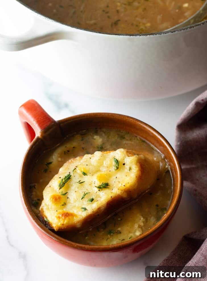 Large red mug with French onion soup, topped with a slice of baguette with melted cheese, ready to be enjoyed.