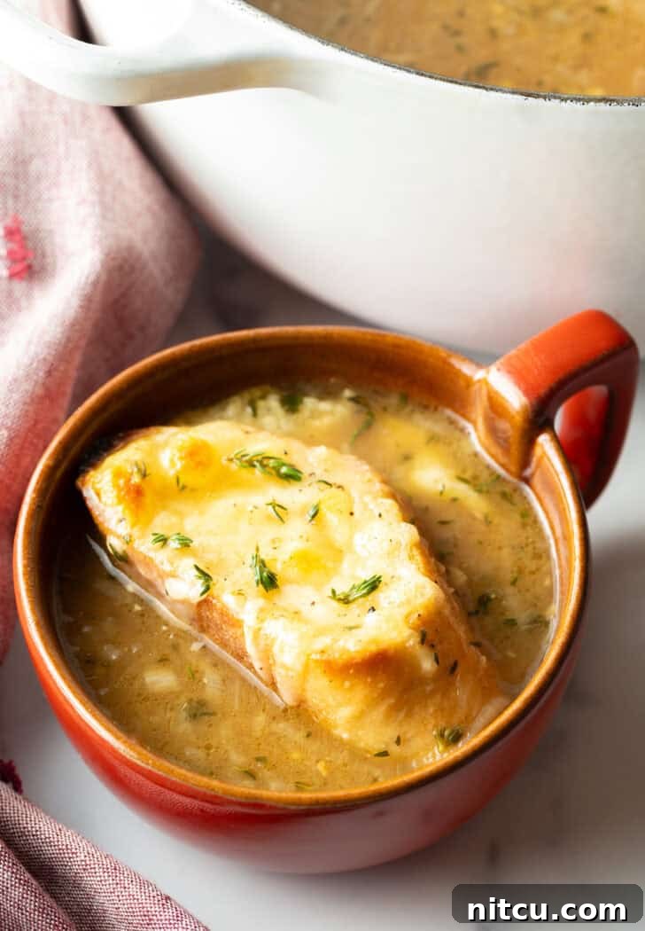 Large red mug with French onion soup, topped with a slice of baguette with melted cheese, ready to be served.