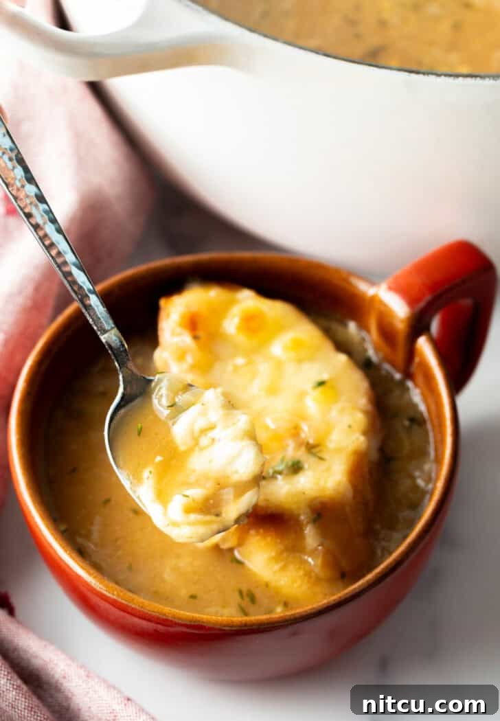 Large red mug with French onion soup, topped with a slice of baguette with melted cheese. A spoon is going in for a scoop.