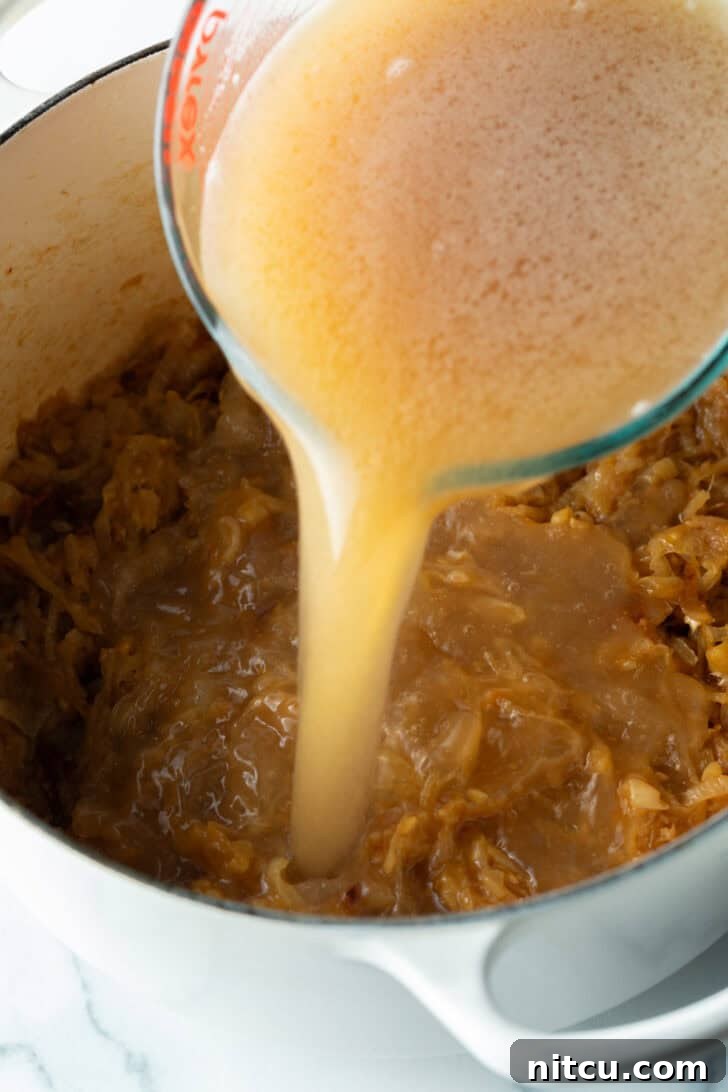 Pouring beef stock from a glass pitcher into the large pot with cooked onions.