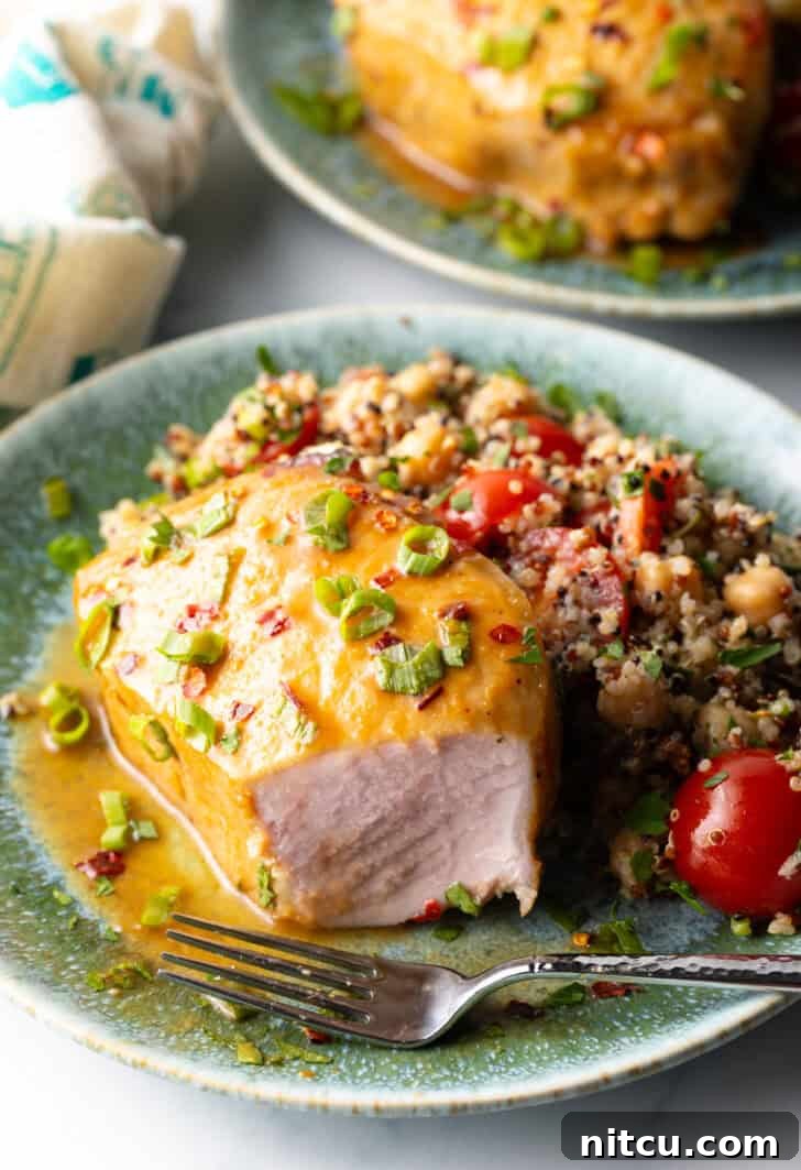 A perfectly baked pork chop, covered with a shiny glaze and fresh chopped scallions, on a plate next to quinoa salad. A fork is strategically placed, and a large portion of the front of the chop has been expertly removed to showcase the tender, juicy interior meat.
