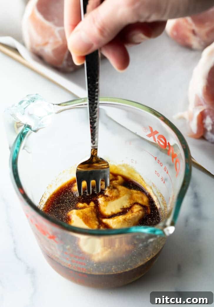 A close-up of a fork stirring together light brown sugar, soy sauce, and Dijon mustard, along with garlic powder, in a clear glass measuring cup, showcasing the beginnings of the rich glaze.