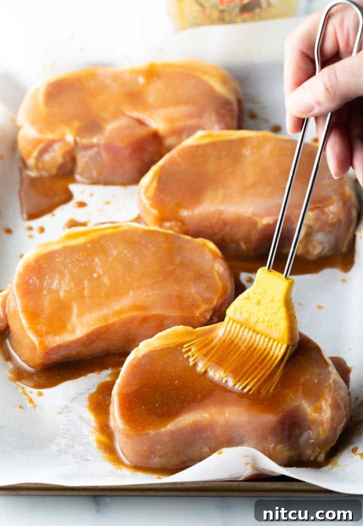 A chef's brush carefully applying the rich, savory-sweet glaze over raw pork chops arranged on a baking sheet, ensuring full coverage before baking.