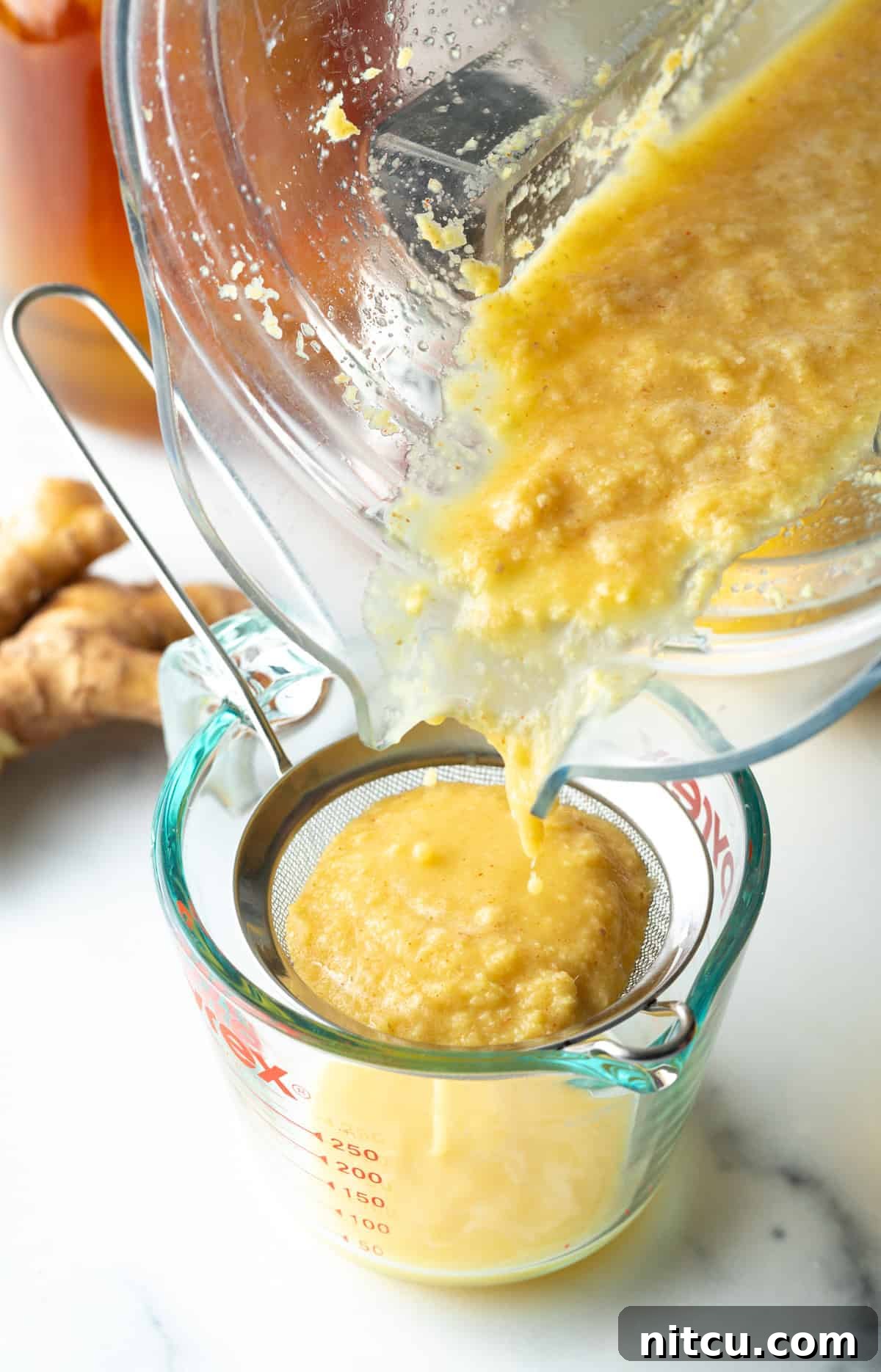 Pouring pureed ginger mixture from a food processor into a sieve set over a glass measuring pitcher, illustrating the straining process.