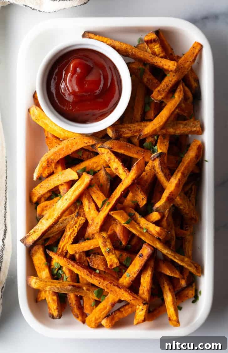Piles of cooked air fryer sweet potatoes on a large serving pan with a white ramekin of ketchup.