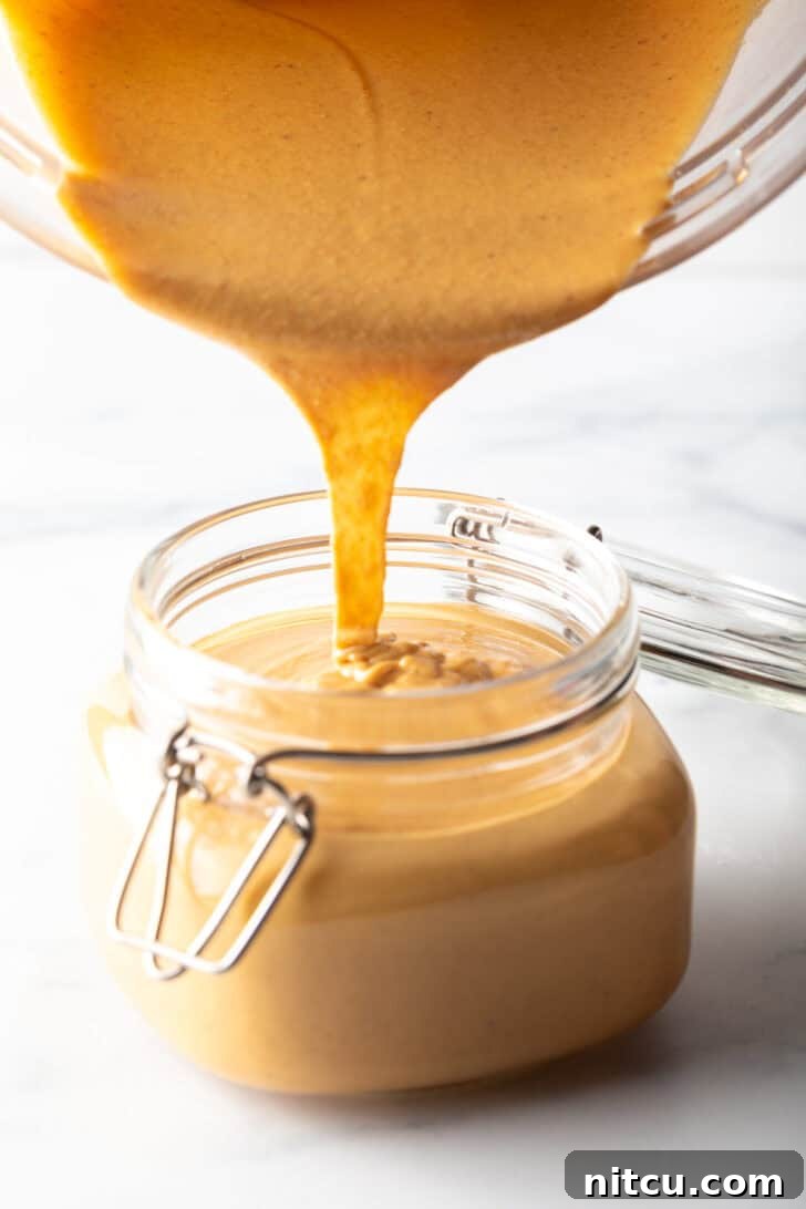 Smooth homemade peanut butter being poured directly from a blender jar into a clear glass storage jar, highlighting its perfectly liquid consistency.
