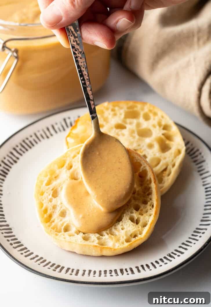 Hand using a spoon to smooth freshly made homemade nut butter onto half of an English muffin, highlighting its delicious texture.
