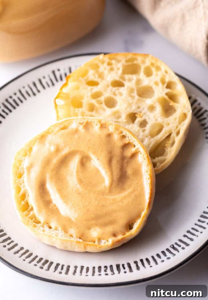 An open English muffin on a plate, with one half generously covered in smooth homemade peanut butter.