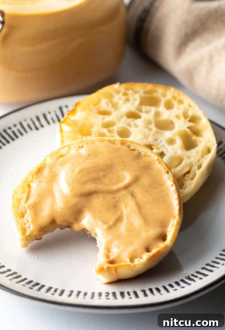 An open English muffin on a plate, one half covered with homemade peanut butter and a bite taken out, showing its creamy texture.