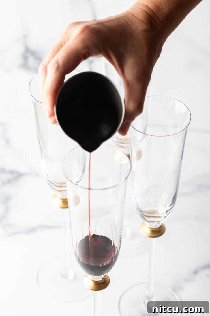 A hand carefully pouring deep red crème de cassis into a champagne flute.