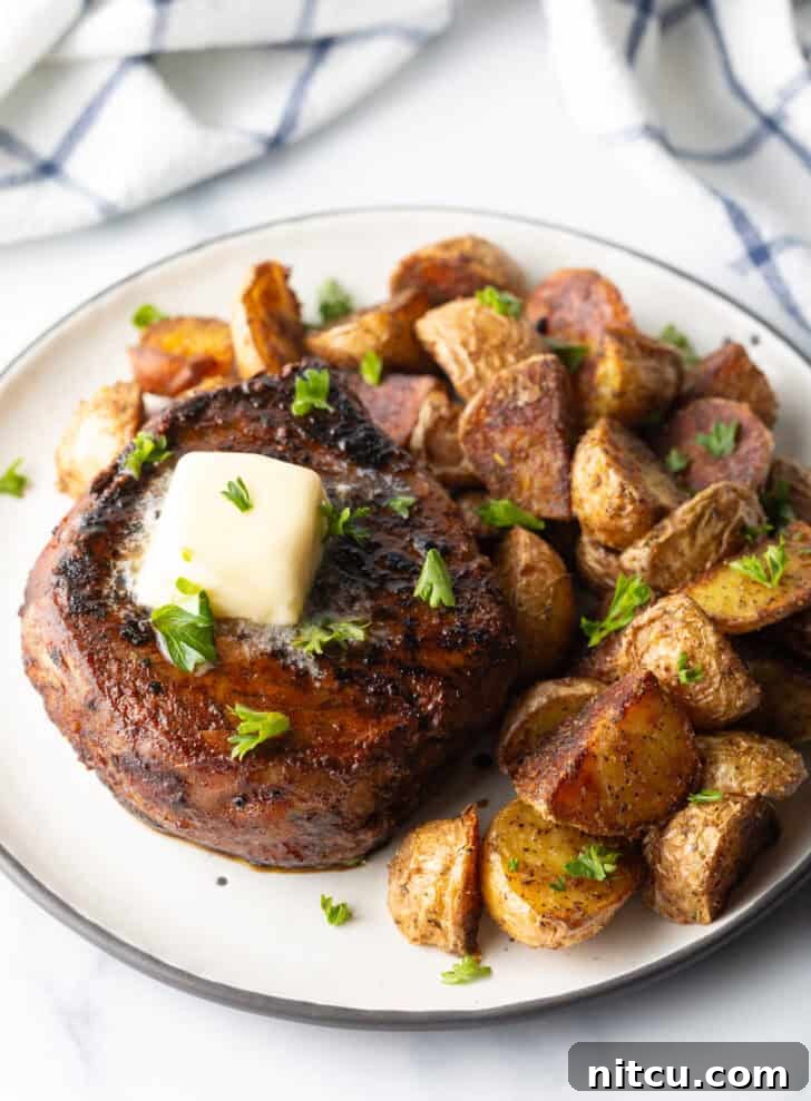 White plate with a reverse sear steak, topped with chopped herbs and a pat of butter. Roasted yellow potato pieces are on the right side of the meat.