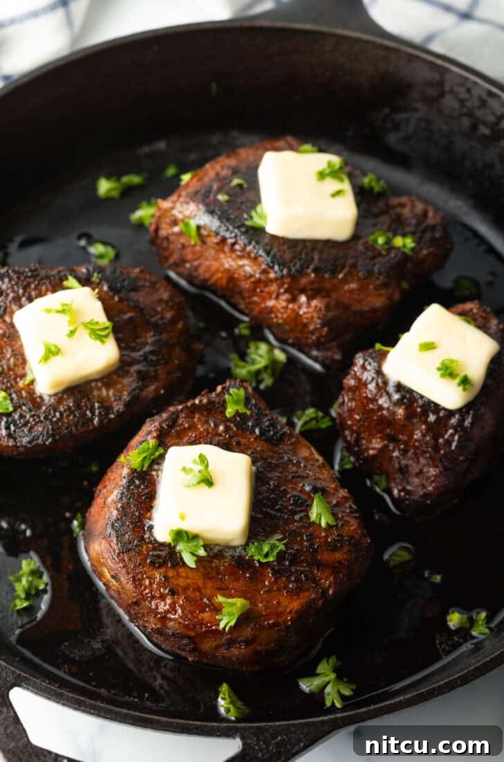 Four filets searing in a cast iron skillet. Each steak is topped with fresh chopped herbs and a pat of butter.