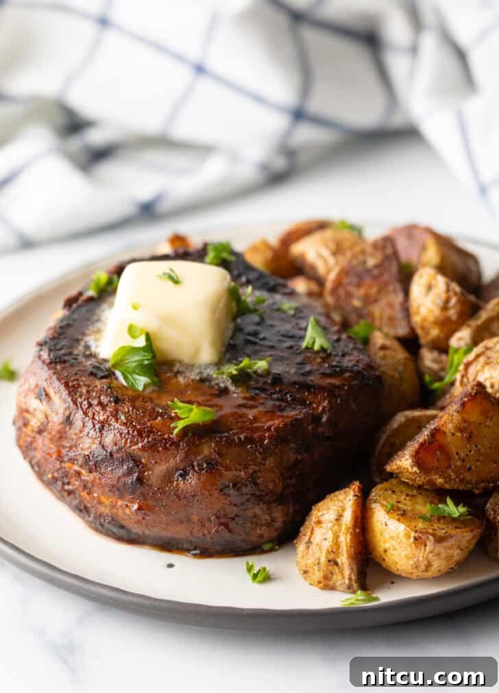 White plate with a reverse sear steak, topped with chopped herbs and a pat of butter. Roasted yellow potato pieces are on the right side of the meat.