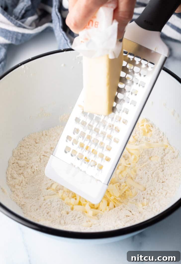 Using a cheese grater to grate butter into a white bowl with dry biscuit ingredients.