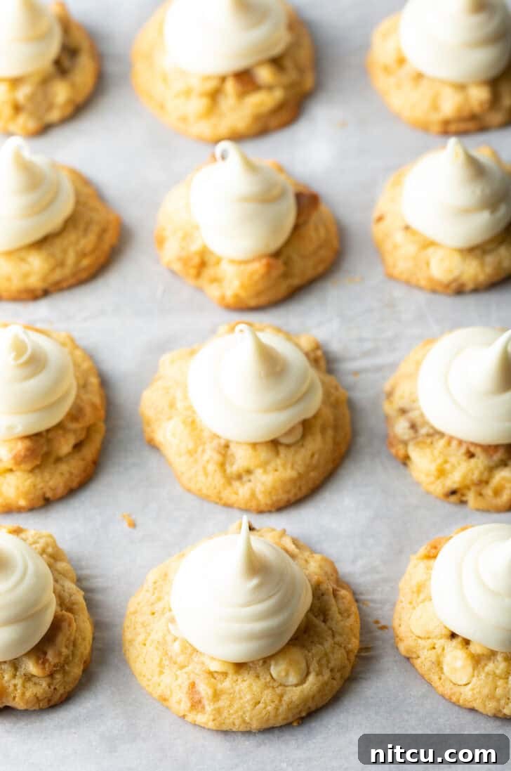 A dozen baked cookies on a baking sheet, each topped with a dollop of vanilla frosting, awaiting final garnishes.