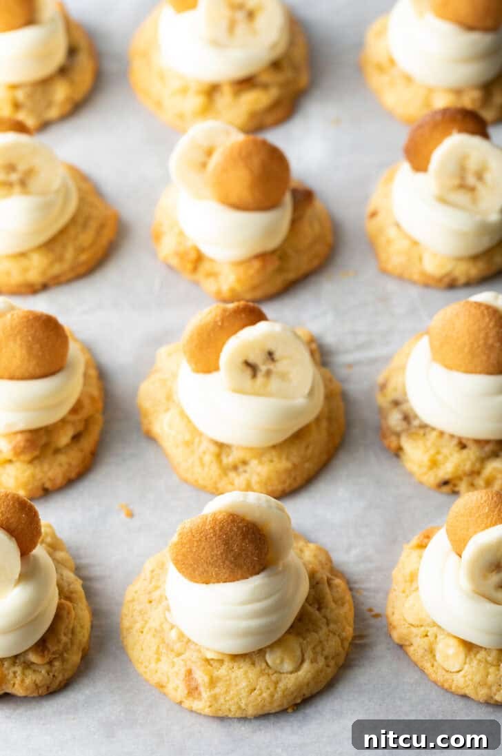 Several banana pudding cookies on a baking sheet, each topped with a dollop of vanilla frosting, a slice of fresh banana, and a Nilla wafer, ready to be enjoyed.