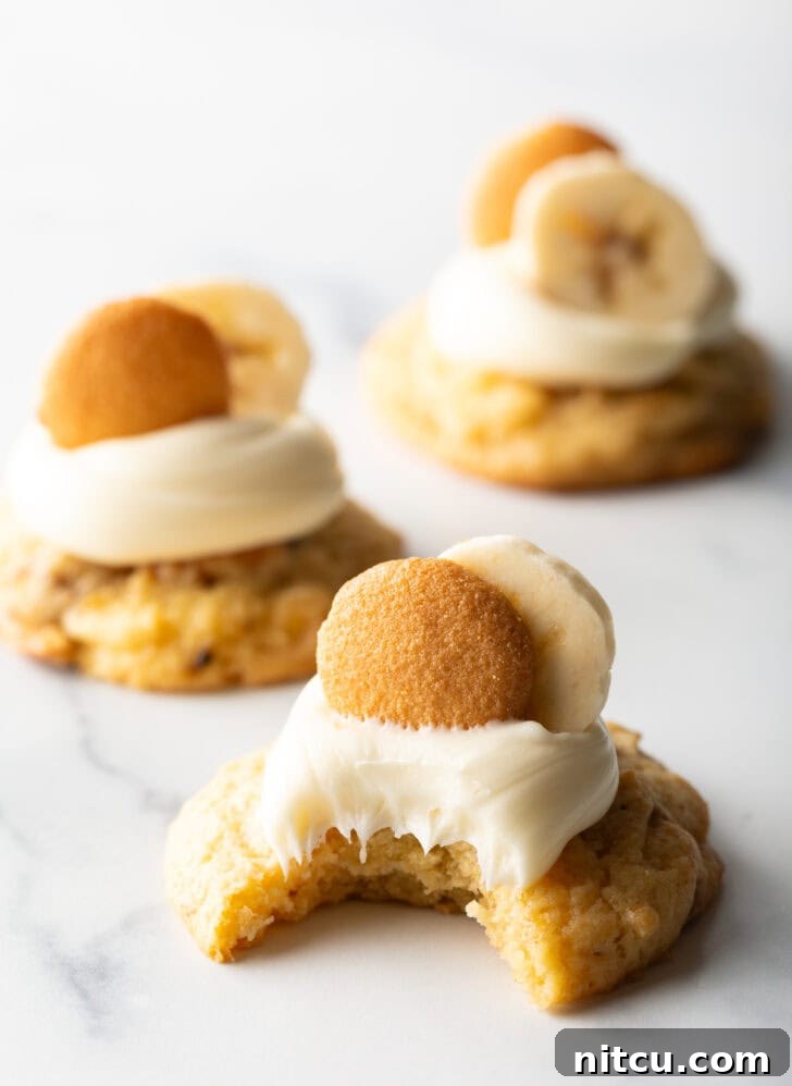 Close view of a banana pudding cookie topped with frosting plus a slice of banana and a Nilla wafer. A bite has been taken out of the cookie, revealing its soft interior.