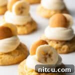 Several banana pudding cookies on a baking sheet, each topped with a dollop of vanilla frosting, a slice of fresh banana, and a Nilla wafer.