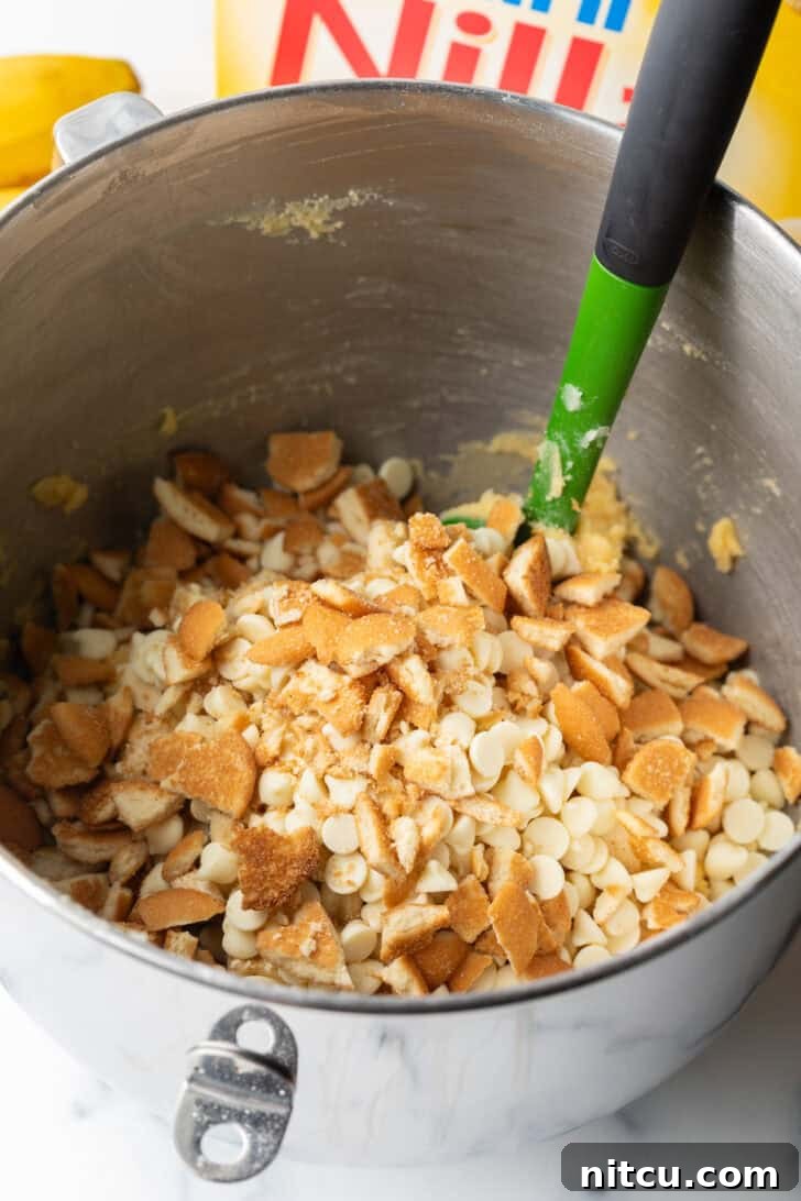 Stirring crumbled Nilla wafers into a metal mixing bowl with cookie ingredients, alongside white chocolate chips and diced banana.