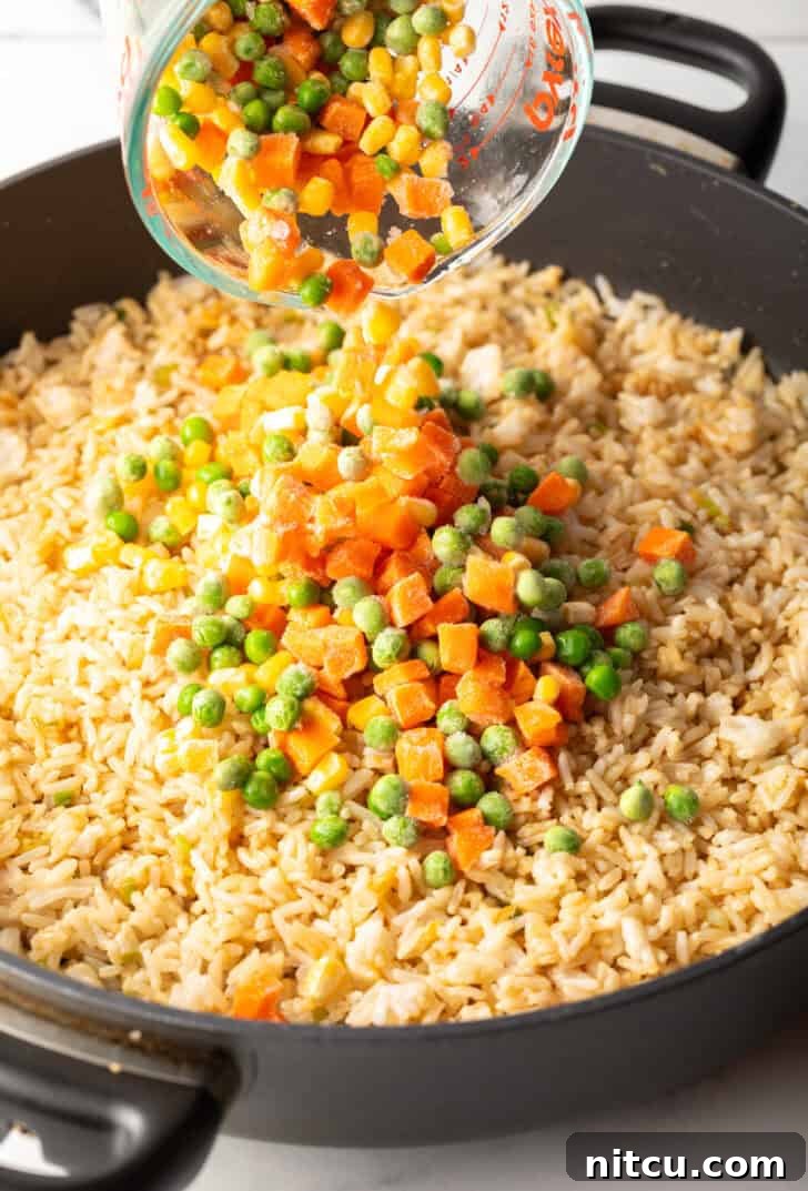 A mix of frozen carrots, peas, and corn kernels being added from a clear glass mixing cup into the rice in a skillet.