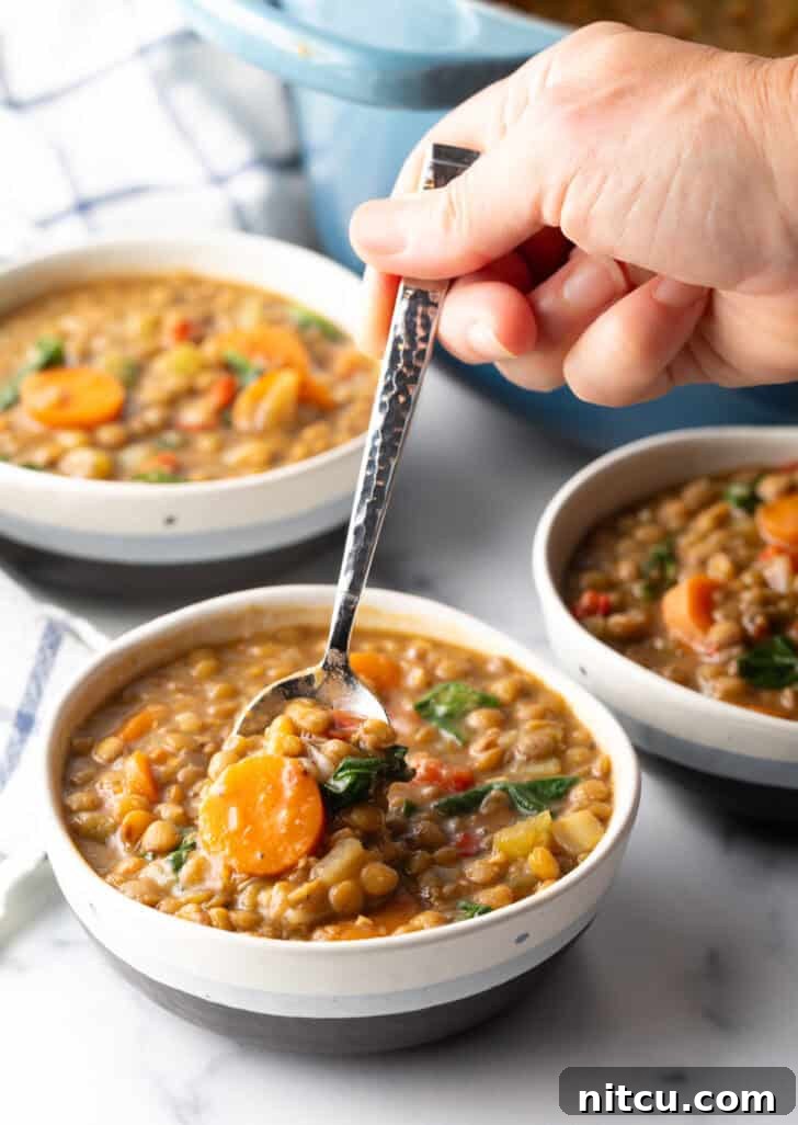 A hand holding a spoon, scooping a generous serving of traditional lentil soup from a white bowl, capturing the inviting texture of the dish.