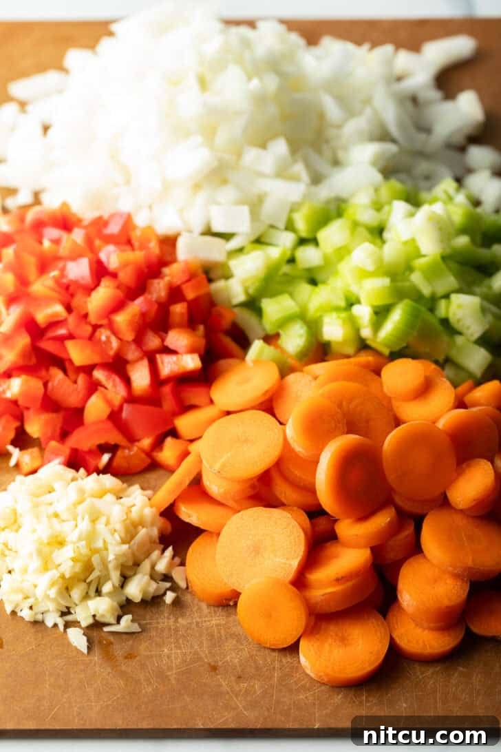 Finely chopped fresh ingredients, including vibrant onion, red peppers, sweet carrots, pungent garlic, and crisp celery, arranged neatly on a rustic wood cutting board, ready for soup preparation.