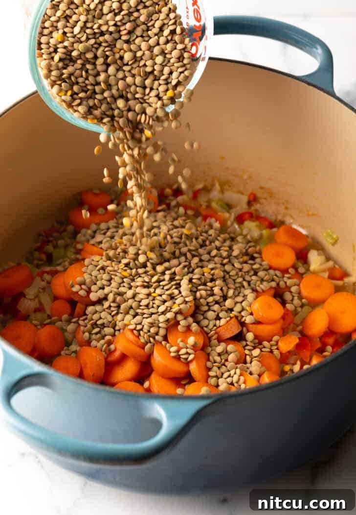 Dried lentils being poured from a clear measuring glass into a large cooking pot already containing sautéed carrots and other vegetables, illustrating a key step in soup preparation.
