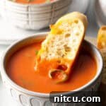 White bowl of tomato soup with a slice of baguette in the soup.