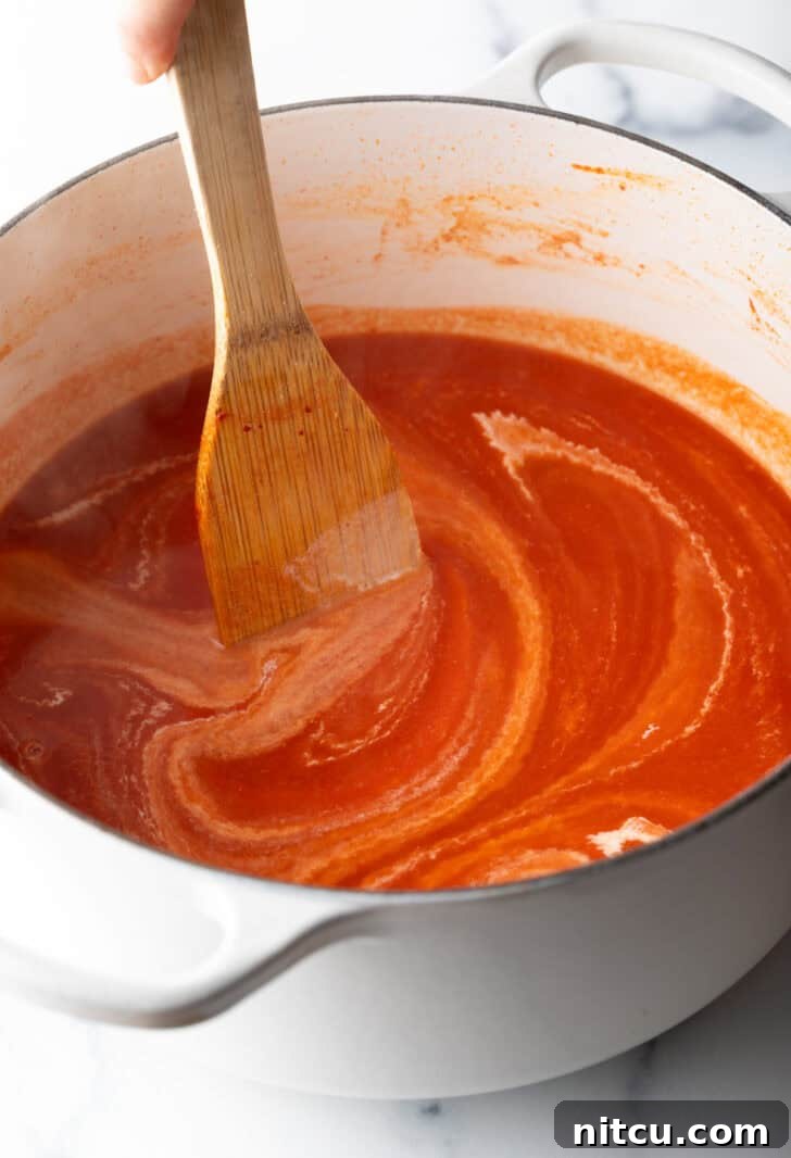 A wooden spatula gently stirring the vibrant red, creamy tomato soup within a cooking pot, creating a swirling motion.