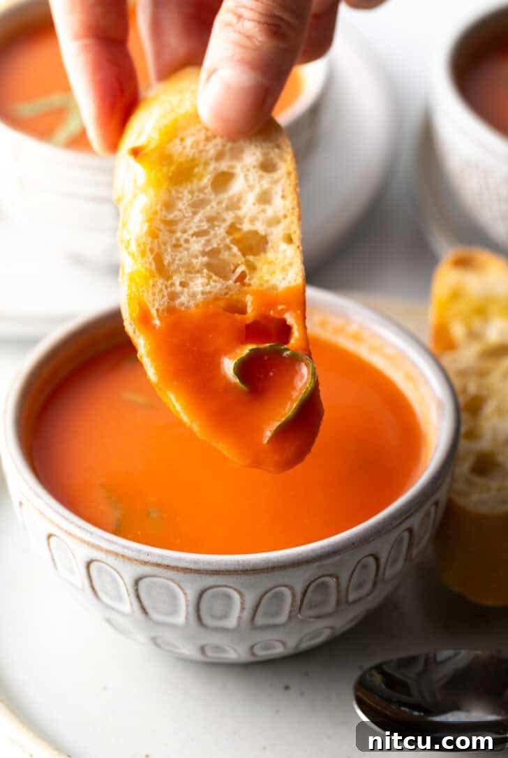 A hand gently dipping a crisp slice of baguette into a bowl of rich, creamy tomato soup, inviting a taste.