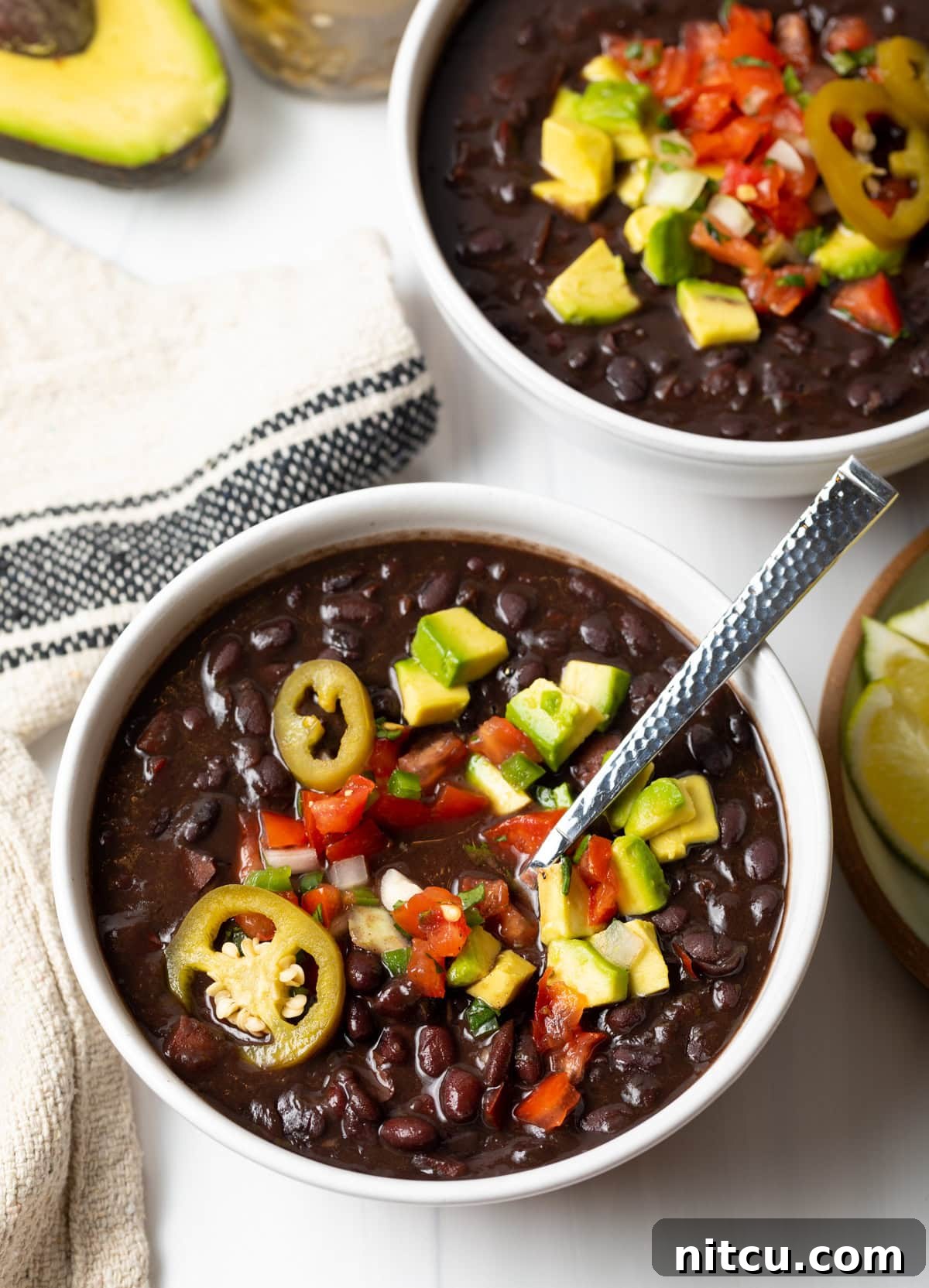 White bowl of black bean soup topped with slices of jalapenos, chopped avocado, and pico de gallo.