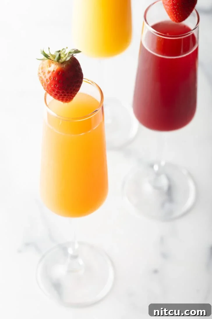 A champagne flute filled with orange mimosa, and another glass filled with a cranberry mimosa. Both glasses are garnished with a whole strawberry on the rim.