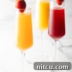 Two champagne flutes filled with orange mimosa, and another glass filled with a cranberry mimosa. Both glasses are garnished with a whole strawberry on the rim.