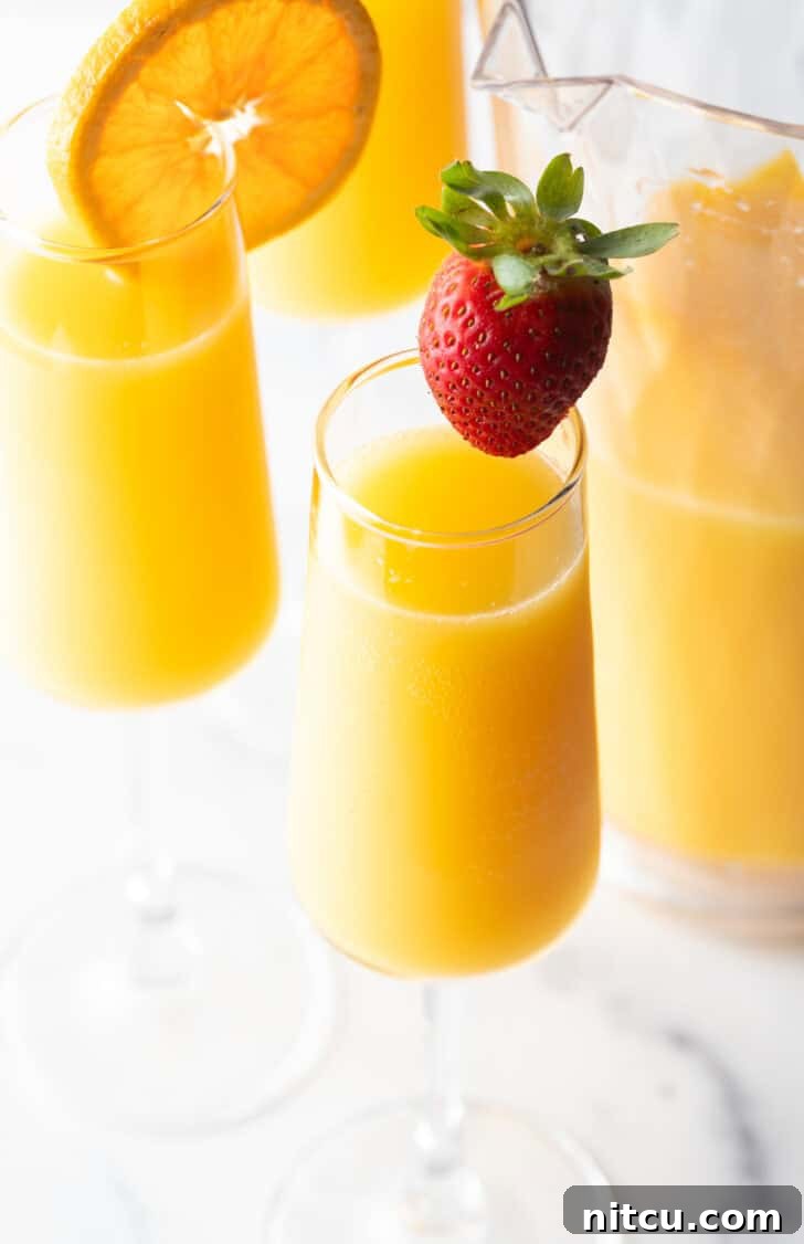 Two glasses of mimosas, one garnished with a large strawberry on the rim, the other garnished with an orange slice on the rim.