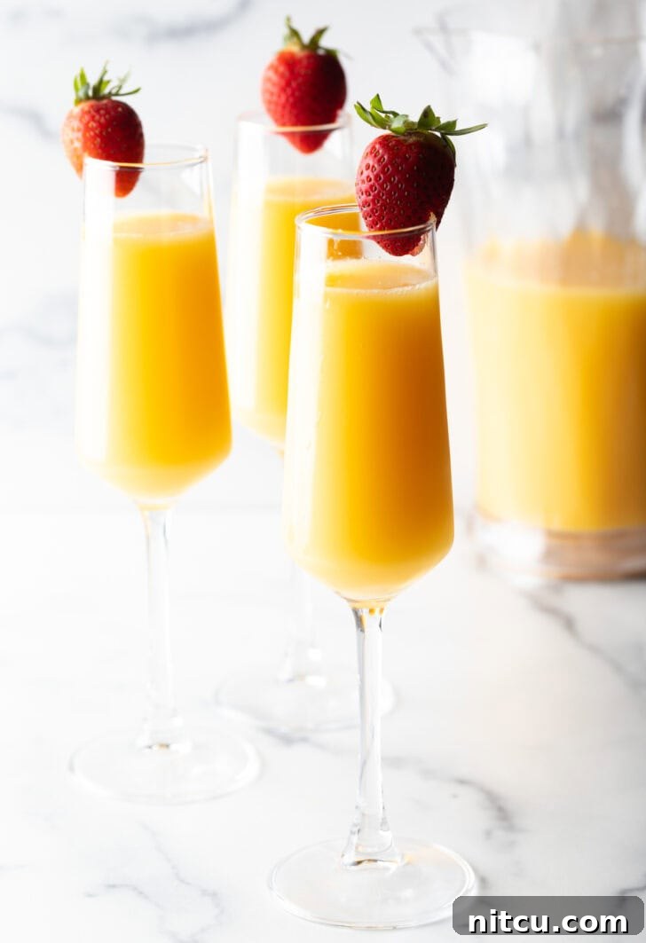 3 champagne glasses full of mimosa and each garnished with a whole strawberry on the rim.