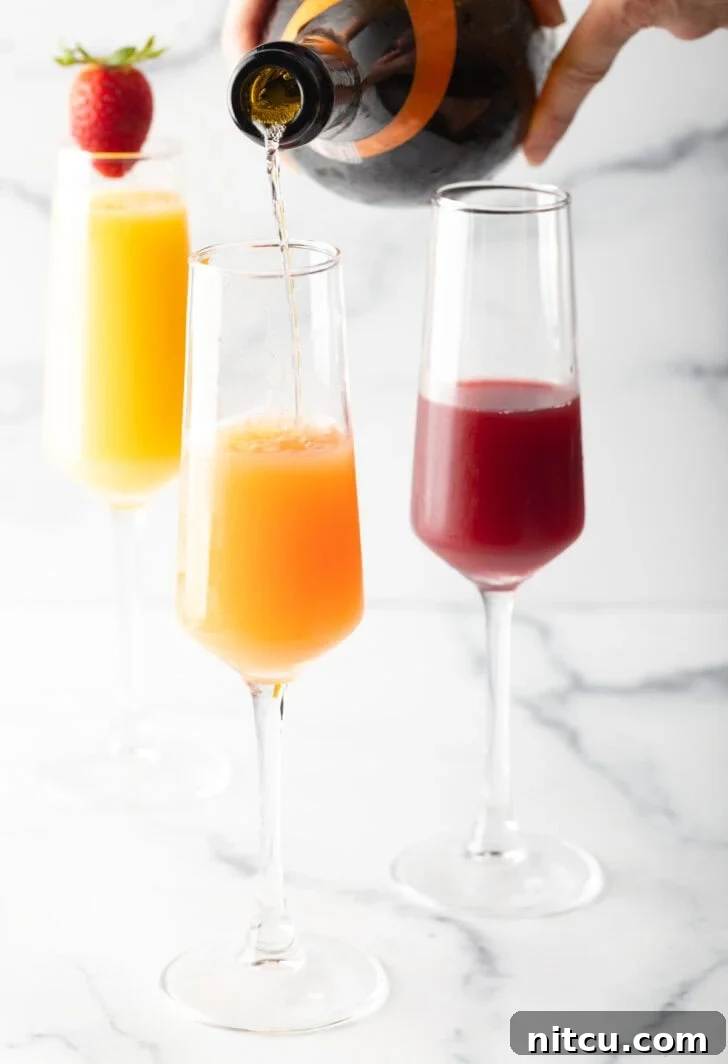 3 champagne glass against a white marble backdrop. The glass in the background is full of orange juice and garnished with a whole strawberry on the rim. In the foreground are a glass half full with cranberry juice, and a glass half full of orange juice with sparkling wine being poured in to the glass.