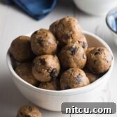 White bowl brimming with energy balls that are speckled with chocolate chips and raisins. One ball is on the table next to the bowl.