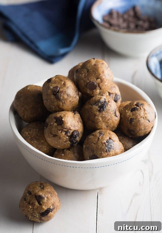 White bowl brimming with energy balls that are speckled with chocolate chips and raisins. One ball is perfectly placed on the table next to the bowl, inviting a taste.