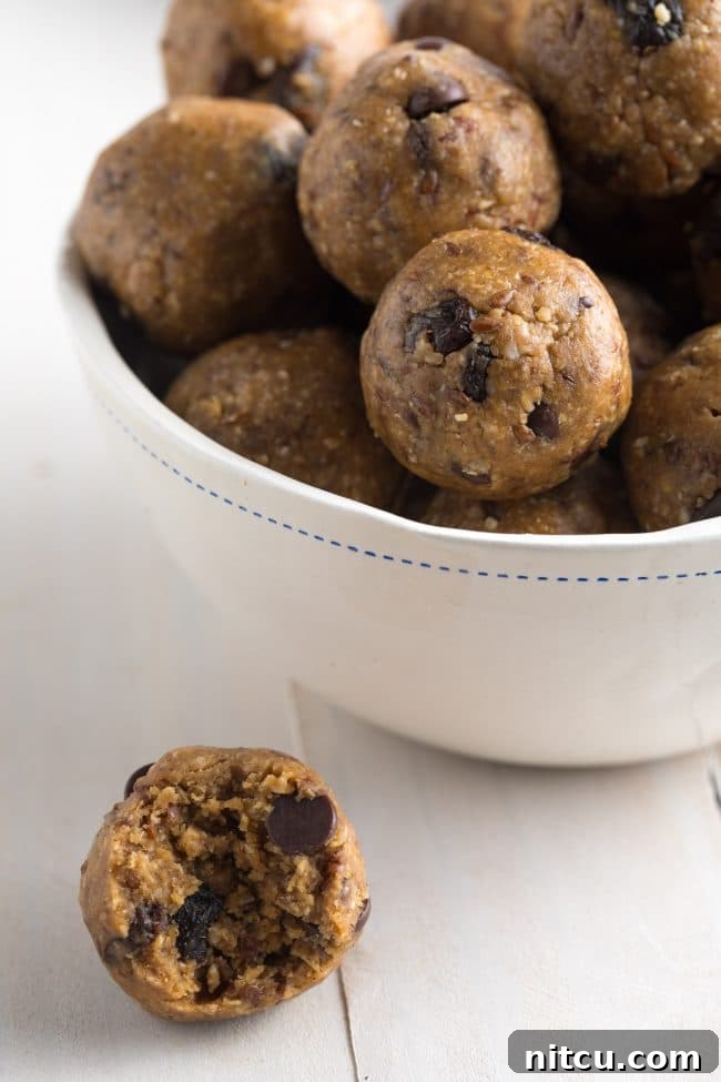 White bowl brimming with energy balls that are speckled with chocolate chips and raisins. One ball with a bite taken out of it is on the table next to the bowl, highlighting its inviting texture.