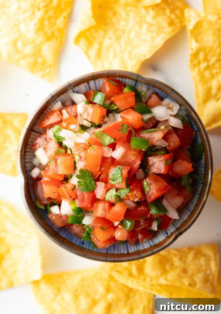 Top down view of a white bowl of fresh pico de gallo with chopped tomatoes, onion, jalapeno, and cilantro. The bowl is surrounded by tortilla chips.