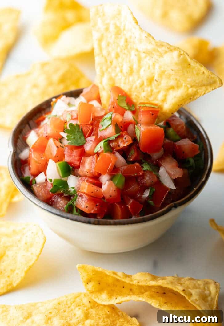 A white bowl of fresh pico de gallo with chopped tomatoes, onion, jalapeno, and cilantro. A tortilla chip is sticking out of the bowl.