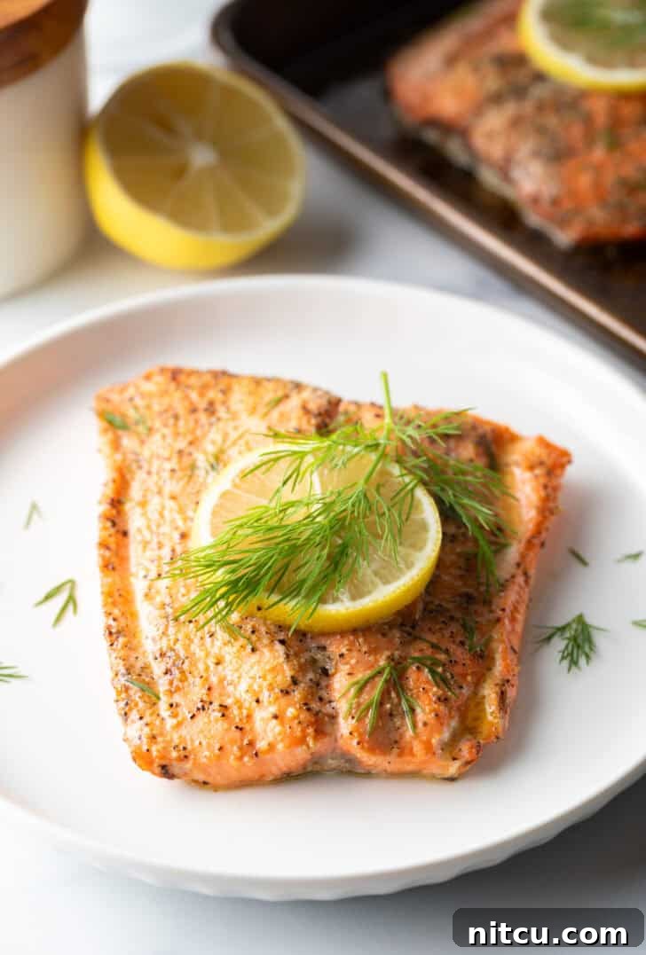 Large filet of salmon topped with a lemon slice and sprigs of fresh dill. The fish is on a white plate.