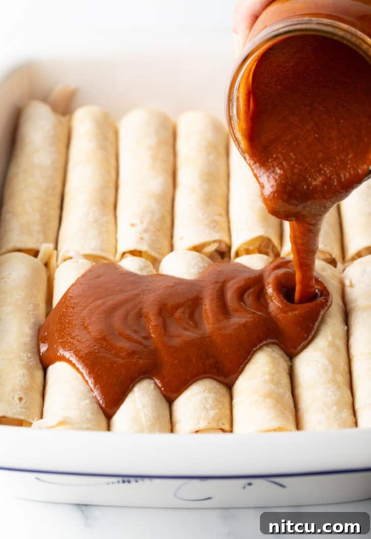 Pouring red sauce from a glass jar over the top of a dozen rolled enchiladas in a white baking dish.
