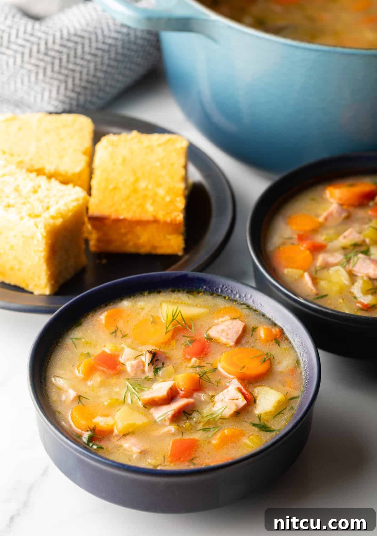 Two black bowls of ham and cabbage soup next to a black plate with 3 squares of cornbread.