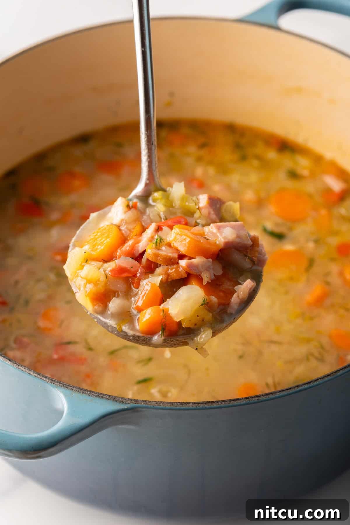 Metal ladle holding a scoopful of cabbage ham soup to camera, held over a blue cooking pot of soup.